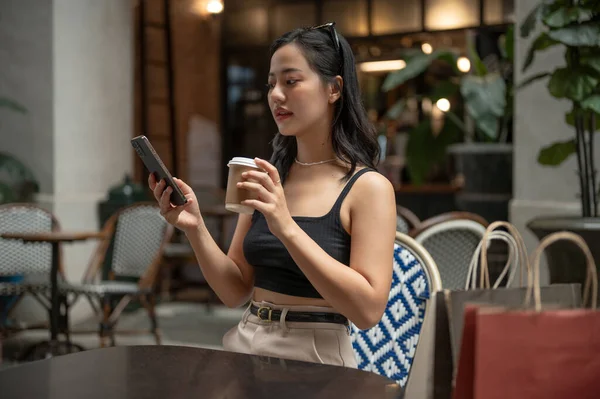 Güzel ve kendine güvenen Asyalı bir kadın kahvesini yudumluyor ve şehirdeki bir kafe veya restoranda dinlenirken akıllı telefonunu kullanıyor. Yaşam tarzı kavramı