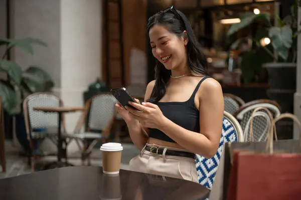 A happy and confident Asian woman in trendy clothes is using her phone, chatting with her friends on her phone while relaxing in a cafe or restaurant in the city. Lifestyle concept