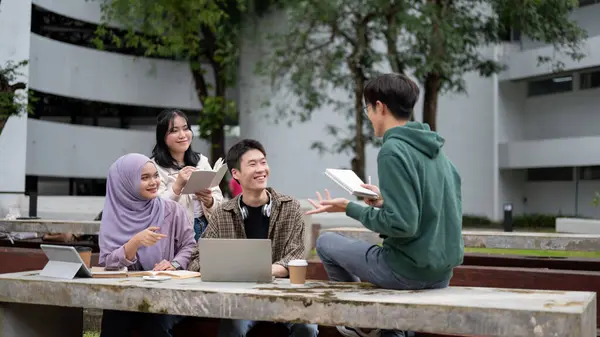 A smart and cheerful young Asian male college student in a green hoodie is helping his friends with math, tutoring and preparing for the exam in a campus park with his friends. Education concept