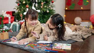 İki sevimli ve mutlu Asyalı genç kız Noel ağacının yanında oturma odasında uzanıyorlar ve birlikte çizim ve boyamanın keyfini çıkarıyorlar. Çocuk eğlencesi, yaratıcılık aktivitesi, Mutlu Noeller.