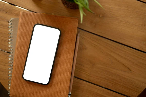 A white-screen smartphone mockup on a book on a wooden table. study table, office desk, minimal workspace top view image