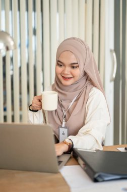 Neşeli ve çekici bir Asyalı-Müslüman iş kadını sabah kahvesini ofisteki bilgisayarında çalışırken içiyor..