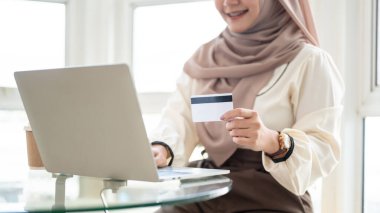 Başörtüsü takan güzel bir Asyalı-Müslüman kadının kesilmiş görüntüsü bir kafede otururken bilgisayarından alışveriş yapıyor, dizüstü bilgisayar kullanıyor ve elinde kredi kartı tutuyor..