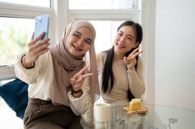 Güzel ve mutlu bir Asyalı-Müslüman kadın bir kafede eğlenirken arkadaşıyla selfie çekiyor..