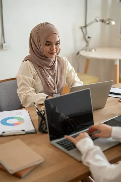 A professional and beautiful millennial Asian-Muslim businesswoman in a hijab is in the meeting with her team, working on her laptop and discussing work with her colleagues.