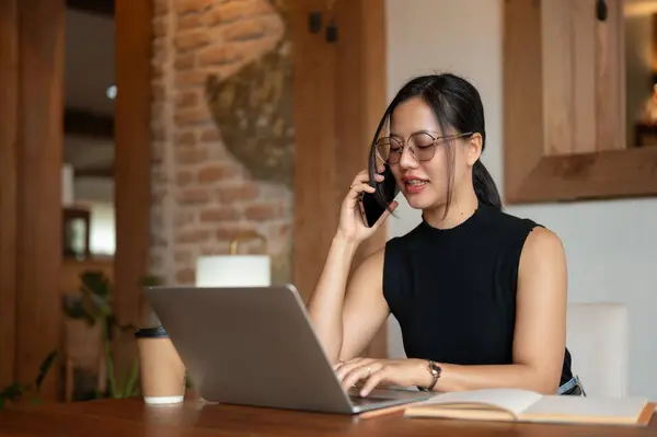 A professional and attractive millennial Asian businesswoman is talking on the phone with her client while working remotely from a coffee shop in the city. City life, digital nomad, freelance