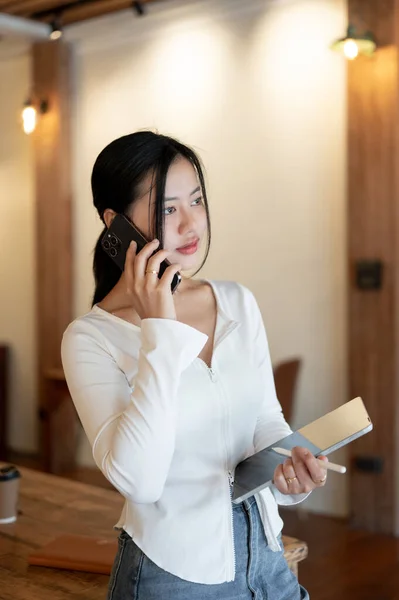 A portrait of a beautiful Asian woman in casual clothes is calling or talking with someone on her phone while standing in a coffee shop.