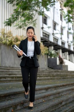 Resmi takım elbiseli, meşgul ve profesyonel Asyalı bir iş kadını binanın önünde merdivenlerden inerken müşterisiyle telefonda konuşuyor..