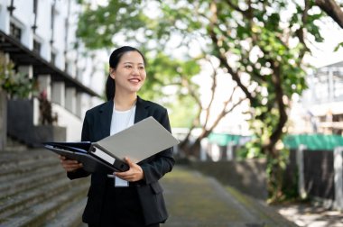 Resmi bir iş elbisesi içinde başarılı ve mutlu bir Asyalı iş kadını elinde klasörlerle binanın dışında kameraya bakmıyor ve hayal kuruyor..