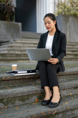 Resmi takım elbiseli, meşgul, konsantre Asyalı bir iş kadını şehirdeki ofis binasının önündeki merdivenlerde otururken dizüstü bilgisayarı üzerinde çalışıyor..