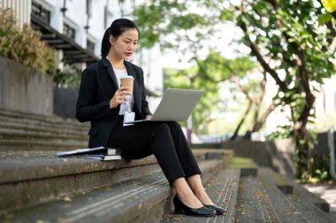 Meşgul, konsantre Asyalı iş kadını resmi bir iş elbisesi içinde laptopunu yudumlarken şehirdeki ofis binasının önündeki merdivenlerde oturuyor..