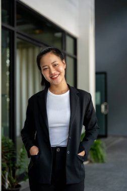 Resmi takım elbiseli güzel ve başarılı bir Asyalı iş kadınının portresi kameraya gülümsüyor ve şirket binasında duruyor..