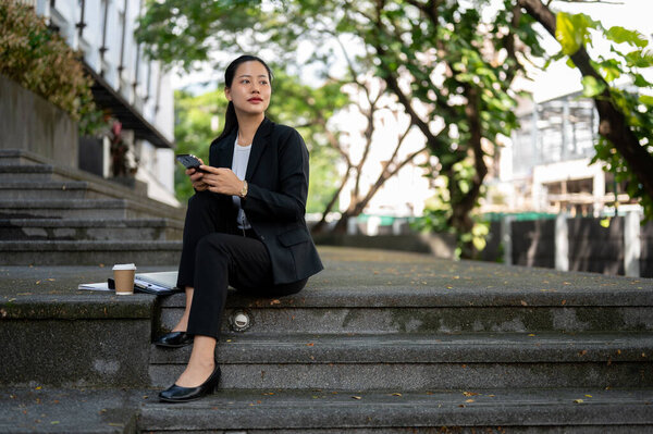 A beautiful millennial Asian businesswoman is chatting on her phone and waiting for someone while sitting on the stairs outdoors in the city.