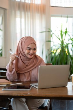 Mutlu ve çekici, tesettürlü genç bir Asyalı-Müslüman kadın bir kahve dükkanında çalışıyor, dizüstü bilgisayarıyla işini yürütüyor..
