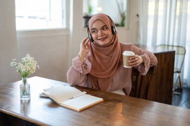Rahatlamış ve mutlu bir Asyalı-Müslüman kadın tesettür ve kulaklık takmış bir kafede sabah kahvesini içerken müzik dinliyor. Yaşam tarzı ve boş zaman kavramları