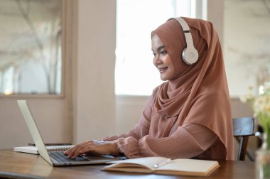 Başörtüsü ve kulaklık takan güzel bir Asyalı-Müslüman kadının yan görüntüsü dizüstü bilgisayarında çalışıyor, online bir toplantı yapıyor, bir kahve dükkanında uzaktan çalışıyor..