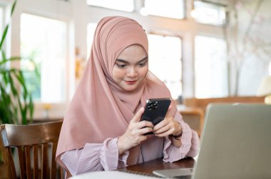 Kafasında tesettür olan mutlu bir Asyalı-Müslüman kadın, kafede uzaktan çalışırken akıllı telefonuyla birilerine mesaj atıyor. İnsanlar ve teknoloji kavramları