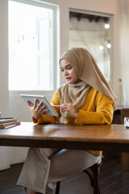 Yoğunlaşmış bir Asyalı-Müslüman kadın dijital tableti üzerinde çalışıyor, internetten bir projeyi kontrol ediyor, şehirdeki bir kafede çalışıyor..