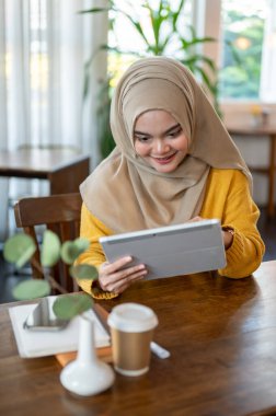 Mutlu ve çekici bir Asyalı-Müslüman kadın dijital tablet kullanıyor, sosyal medyada kayıyor veya güzel bir kafede dinlenirken çevrimiçi videolar izliyor..