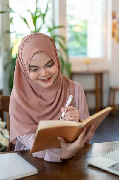 A portrait of an attractive Asian-Muslim woman wearing a hijab is reading a book or keeping her diary while sitting in a coffee shop. People and lifestyle concepts