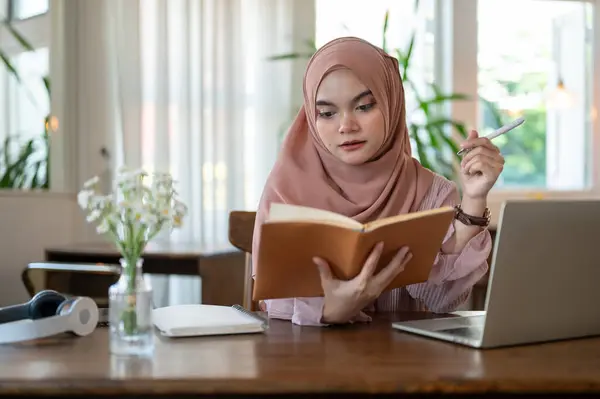 A beautiful and concentrated young Asian-Muslim woman wearing a hijab is reading a book while working remotely at a coffee shop. People and lifestyle concepts