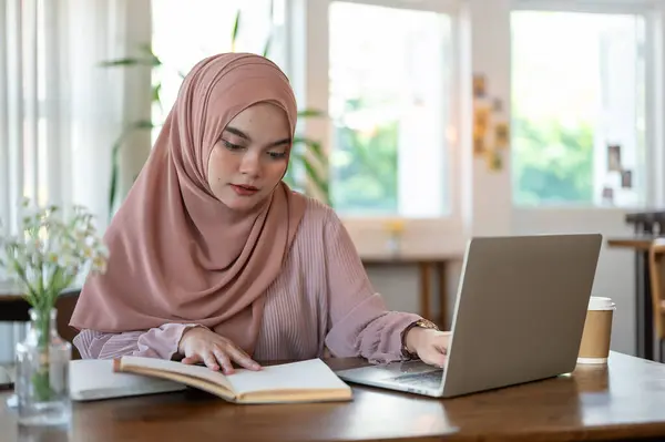 A concentrated and thoughtful young Asian-Muslim woman wearing a hijab is reading a book and working on her tasks on her laptop in a cafe.