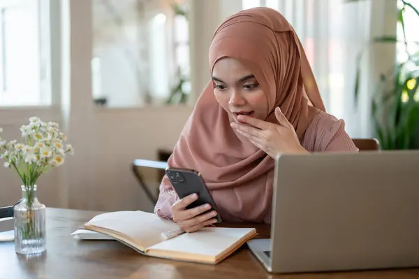 stock image An excited Asian-Muslim woman is looking at her smartphone screen with a surprising face, covering her mouth, shocked with a good news or an unexpected message while sitting at a table in a cafe.