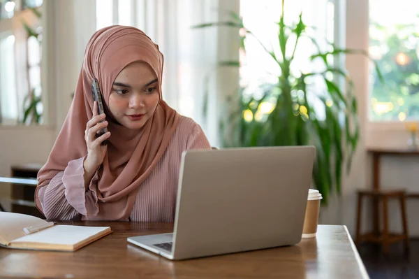 A concentrated Asian-Muslim woman is looking at some details on her laptop screen while talking on the phone with someone, working remotely at a coffee shop.