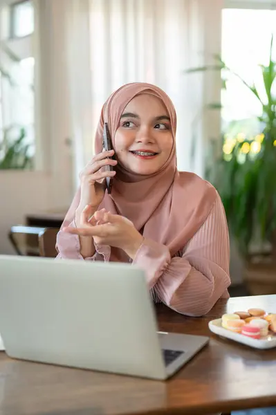 An attractive and cheerful young Asian-Muslim woman is calling someone or talking on the phone with someone while working remotely at a coffee shop. People and technology concepts