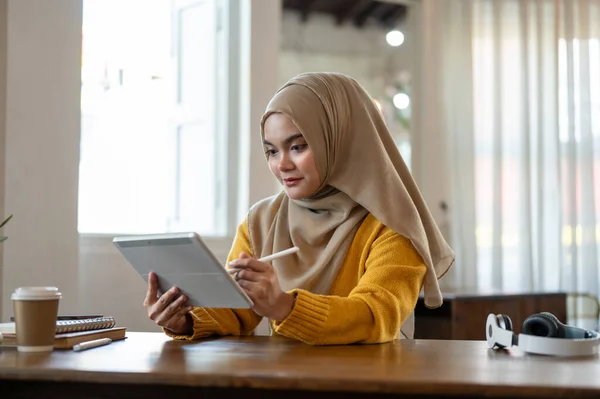 A beautiful Asian-Muslim woman in a hijab is using a digital tablet to manage her work while sitting in a coffee shop. People and wireless technology concepts