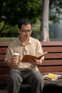 Gözlüklü mutlu bir emekli Asyalı adam yeşil bir parktaki bankta dinlenirken kitap okuyor ve sabah kahvesini içiyor. Mutlu emeklilik yaşam tarzı konsepti