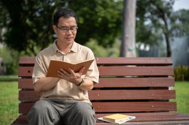 Mutlu bir emekli Asyalı adam yeşil bir parkta bir bankta dinlenirken kitap okuyor. Mutlu emeklilik yaşam tarzı konsepti