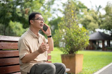 Yaşlı, emekli Asyalı bir adamın, parkta bir bankta dinlenirken kızıyla telefonda konuşurken görüntüsü. Yaşlılar ve teknoloji kavramları