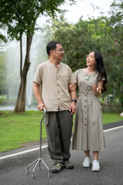 A happy senior Asian dad with a walking stick is talking with his lovely daughter while walking in the green park daughter. Happy family, caregiver, granddaughter and grandfather, dad and daughter