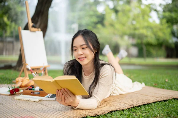 An attractive and happy young Asian woman is reading a book while laying on a picnic mat, enjoying picnicking in a green park on the weekend.