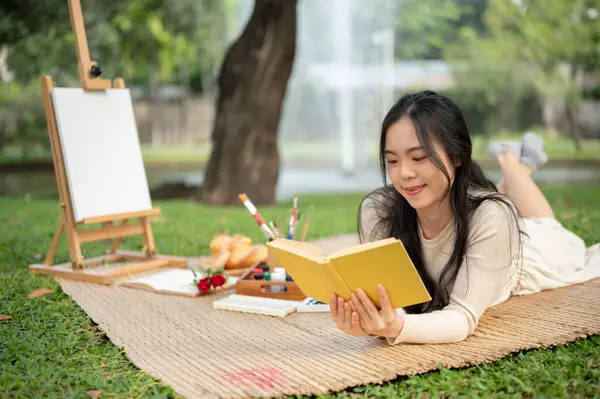 An attractive and happy young Asian woman is reading a book while laying on a picnic mat, enjoying picnicking in a green park on the weekend. Lifestyle concept