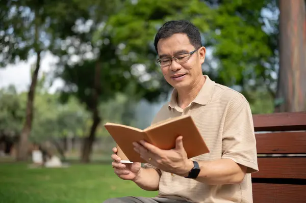 A happy senior retired Asian man wearing eyeglasses is reading a book and having morning coffee while relaxing on a bench in a green park. Happy retirement lifestyle concept