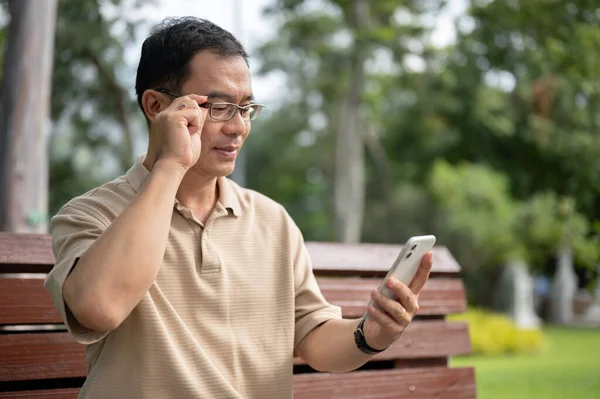A happy retired Asian man adjusting his eyeglasses while reading messages on his smartphone while relaxing on a bench in a park. Old people with technology concepts