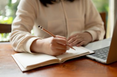 Asyalı bir üniversite öğrencisinin yakın plan fotoğrafı ev ödevi yapıyor, dizüstü bilgisayarı üzerinde çalışıyor ve kapalı bir masada defterine notlar alıyor..