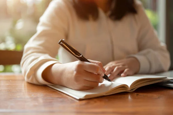 Cropped image of an Asian woman in casual clothes writing something in her book, making lists, taking notes, or keeping her diary at a table indoors.