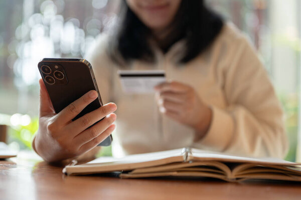Cropped image of a happy and satisfied Asian woman enjoys shopping online on her smartphone, holding a smartphone and a credit card while sitting at a table indoors.