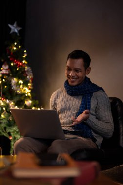 Rahat bir süveter giyen neşeli bir Asyalı, bir Noel gecesi karanlık bir oturma odasında internetten Noel 'i kutlarken dizüstü bilgisayarıyla ailesiyle konuşmaktan zevk alıyor..