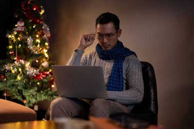 Odaklanmış ve stresli Asyalı bir adam Noel gecesi oturma odasında dizüstü bilgisayarı üzerinde çalışıyor, Noel tatilinde meşgul bir işadamı, Noel 'de evden çalışıyor..