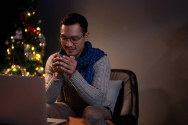 Rahat ve sakin bir Asyalı adam, rahat bir süveter ve eşarp içinde, karanlık bir oturma odasında, bir Noel gecesinde masasında kahvesinin tadını çıkarıyor..