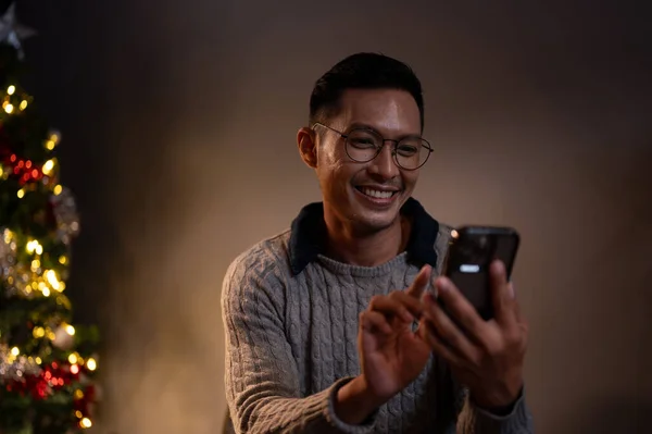 A cheerful Asian man in a cosy sweater is chatting with his friends or sending Christmas messages to his family while relaxing in his living room on a Christmas night.