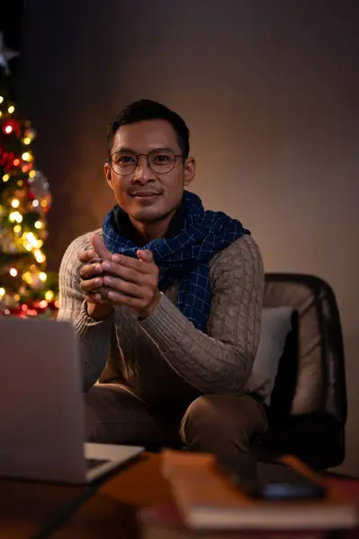 A handsome and happy Asian man in a cosy sweater and scarf is holding a coffee cup while sitting in a dark living room on Christmas night, celebrating Christmas at home alone.
