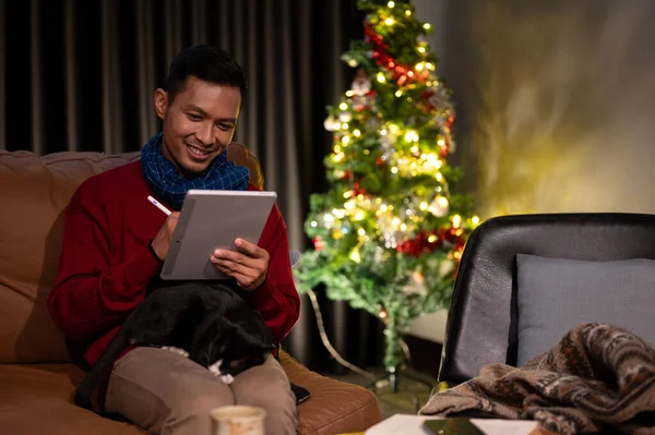 A happy Asian man in cosy clothes is sitting on a sofa and using his tablet while his cat is sleeping on his lap, celebrating Christmas night at home with his cat.