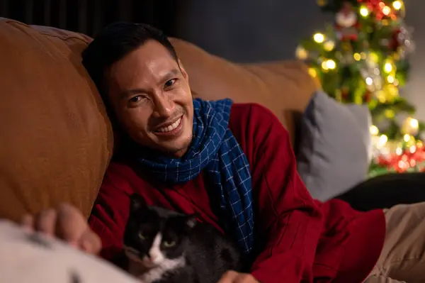 A happy Asian man is lying on a sofa with his lovely cat, celebrating Christmas night at home together. Domestic cat and owner at home. Pet concept