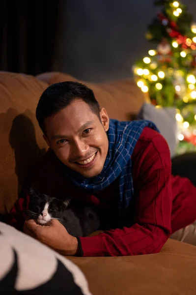 A happy Asian man is lying on a sofa with his lovely cat, celebrating Christmas night at home together. Domestic cat and owner at home. Pet concept