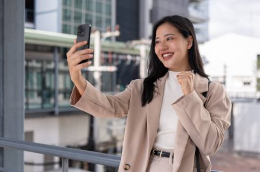 Çekici ve neşeli Asyalı bir iş kadını bir video görüşmesinde konuşuyor ya da şehirdeki hava yürüyüşünde akıllı telefonuyla selfie çekiyor. şehir hayatı konsepti